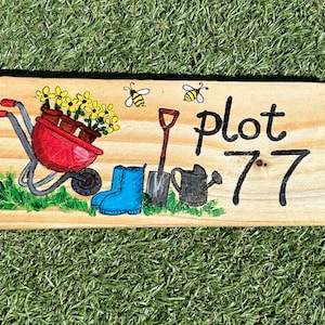May include: A wooden sign with a hand-painted design of a wheelbarrow, gardening tools, and flowers. The sign reads "plot 77".