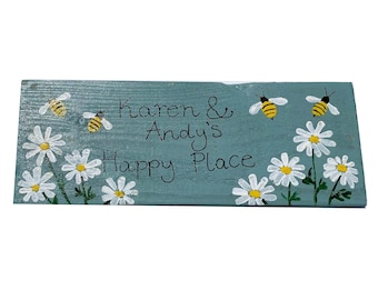 May include: A light blue wooden sign with the text "Karen & Andy's Happy Place" painted in black. The sign is decorated with white daisies and yellow and black bees.