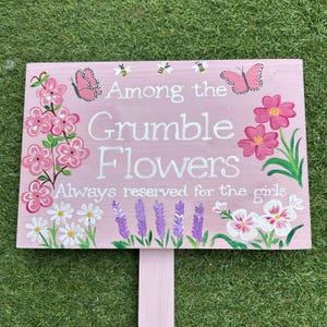 May include: A pink wooden sign with the text "Among the Grumble Flowers Always reserved for the girls." The sign is decorated with painted flowers, butterflies, and bees.