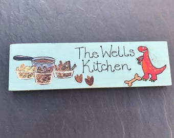 May include: A light blue wooden sign with the text "The Wells Kitchen" in black paint. The sign also features a red dinosaur, a bone, and a few bowls of food.