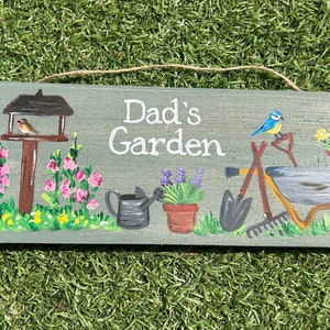 May include: A grey wooden sign with the words "Dad's Garden" painted in white. The sign is decorated with painted images of a bird feeder, flowers, a watering can, a wheelbarrow, and gardening tools.