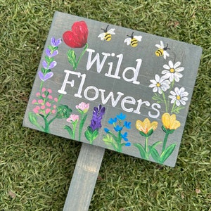May include: A hand-painted wooden sign displaying the words "Wild Flowers" in white. The sign is adorned with colourful flowers, bees, and a red heart. The sign is mounted on a wooden stake, suitable for a garden.