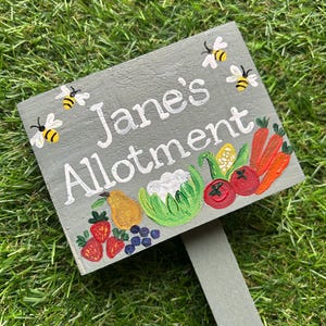 May include: A grey wooden sign with the text "Jane's Allotment" painted in white. The sign is decorated with painted bees and vegetables, including strawberries, blueberries, a pear, cauliflower, corn, tomatoes, and carrots.
