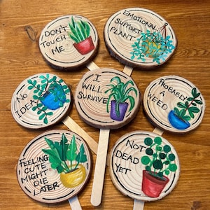 May include: Eight wooden plant markers with painted plants and humorous sayings. The sayings include "No Idea", "Don't Touch Me", "Feeling Cute Might Die Later", "I Will Survive", "Emotional Support Plant", "Probably a Weed", "Not Dead Yet".