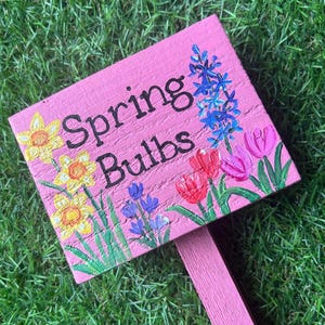 May include: Pink wooden sign with the words "Spring Bulbs" painted in black. The sign is decorated with painted flowers, including daffodils, tulips, and hyacinths.