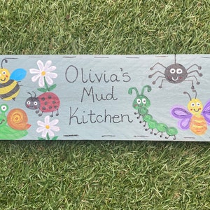 May include: A blue wooden sign with the text "Olivia's Mud Kitchen" painted in black. The sign is decorated with painted images of a bee, a snail, a ladybug, a caterpillar, a butterfly, and a spider.