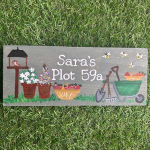 May include: A hand-painted wooden sign with the text "Sara's Plot 59a" in white. The sign features a birdhouse with a red bird, two flower pots with white and purple flowers, a basket of red strawberries, a wheelbarrow, a shovel, and a rake. There are also yellow bees flying around the sign.