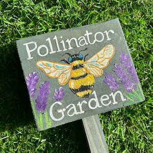 May include: A black wooden sign with white text that reads "Pollinator Garden". A painted bee with yellow and black stripes and blue eyes is in the center of the sign. Purple flowers are painted around the bee.
