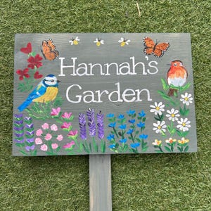 May include: A grey wooden garden sign with the text "Hannah's Garden" in white. The sign is decorated with painted flowers, butterflies, bees, and a blue tit and robin. The sign is mounted on a grey post.