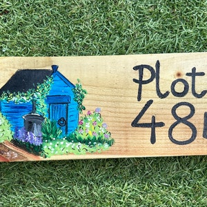 May include: A wooden sign with a hand-painted blue house with a garden and the text "Plot 48b" in black.
