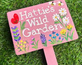 May include: A pink wooden sign with the text "Hattie's Wild Garden" painted in white. The sign is decorated with painted flowers and bees.