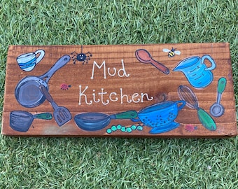 May include: A wooden sign with the words "Mud Kitchen" in white script, surrounded by painted kitchen utensils and insects. The sign features a cup, measuring cup, spoon, and other cooking tools in blue, gray, and green.