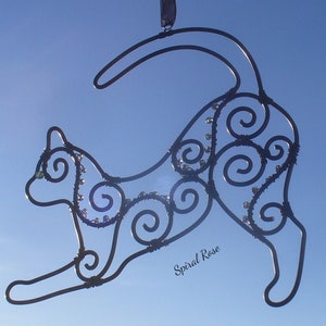 May include: A black metal cat sculpture with a swirling design and small beads. The sculpture is hanging from a string against a blue sky.