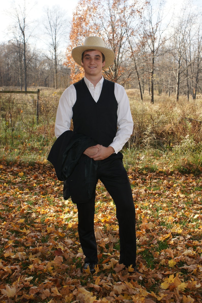 Puede incluir: Un hombre con una camisa blanca, un chaleco negro y pantalones negros est&aacute; de pie en un campo de hojas ca&iacute;das. Lleva un sombrero de paja y tiene una expresi&oacute;n seria en su rostro.