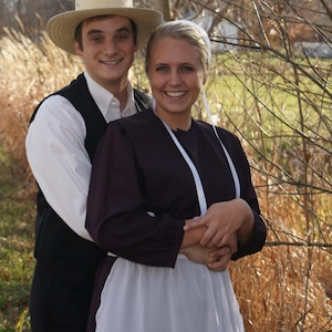 Puede incluir: Una pareja vestida con ropa tradicional Amish. El hombre lleva un sombrero de paja y una camisa blanca con un chaleco. La mujer lleva un vestido largo y oscuro con un delantal blanco y una cinta blanca alrededor del cuello.