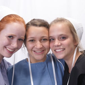 May include: Three young women wearing white bonnets. The woman on the left is wearing a blue dress. The woman in the middle is wearing a light blue dress. The woman on the right is wearing a black dress.