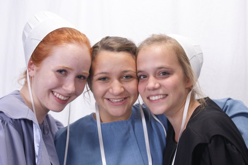 May include: Three young women wearing white bonnets. The woman on the left is wearing a blue dress. The woman in the middle is wearing a light blue dress. The woman on the right is wearing a black dress.