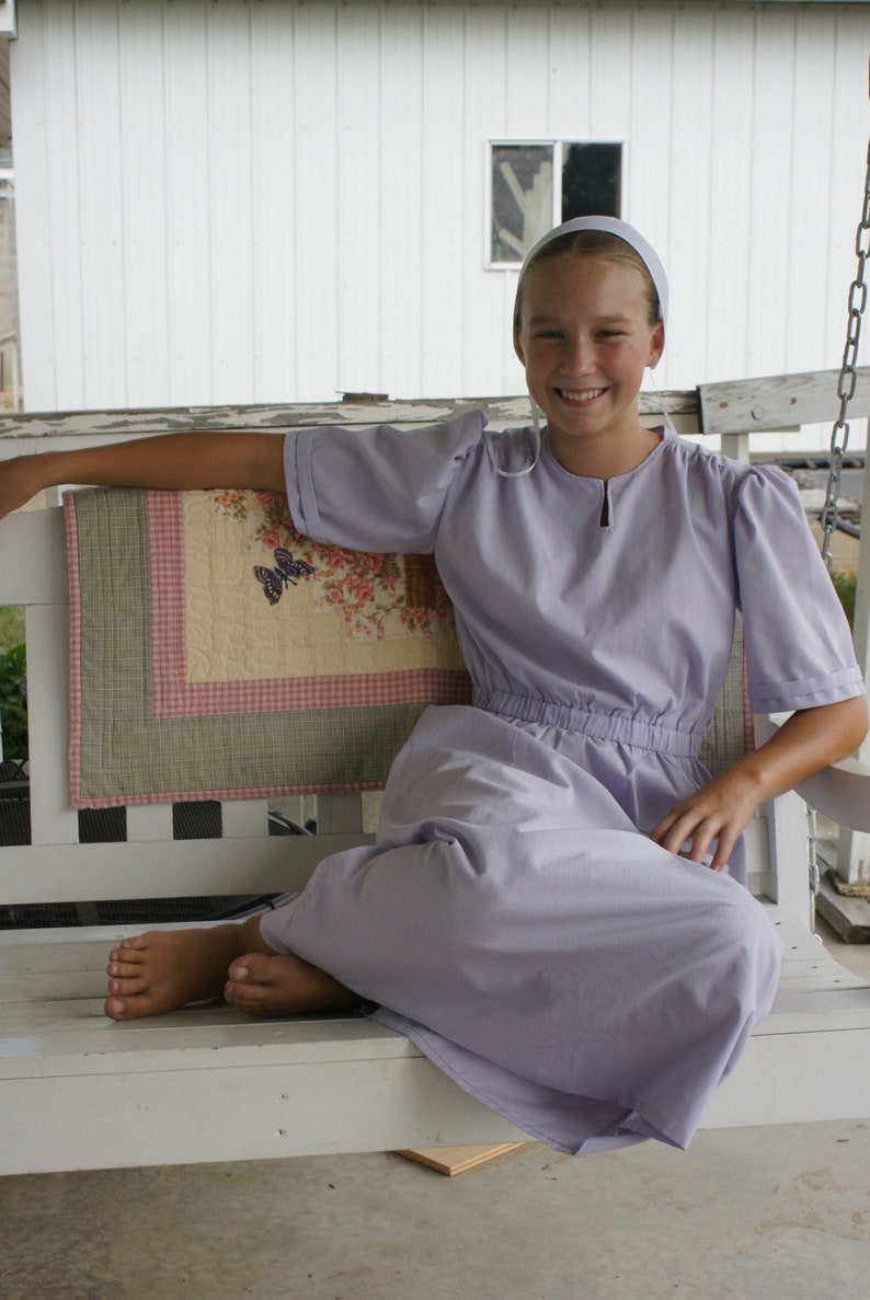 May include: A young person wearing a light purple dress and a white cap sits on a white wooden swing. The person is smiling and looking at the camera. The swing is on a porch with a white wooden railing and a quilt on the back.