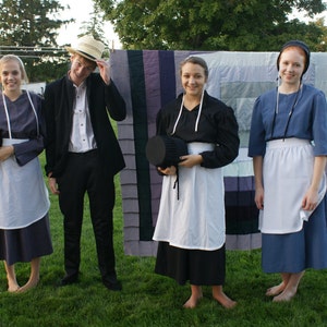 May include: Four people, two women and two men, stand in front of a colorful quilt. The women are wearing long skirts and aprons, and the men are wearing suits and hats. The quilt is made of many different colors and patterns.