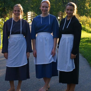 May include: Three women wearing long, dark dresses with white aprons and white head coverings. The women are standing in a grassy area with a wooden fence in the background.