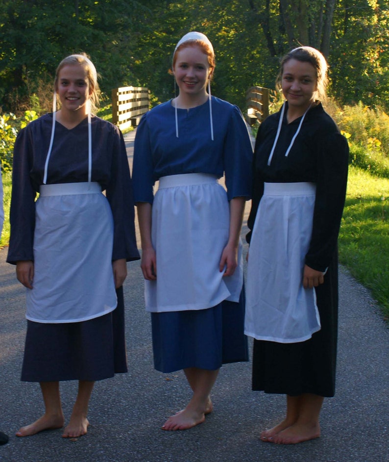 May include: Three women wearing long, dark dresses with white aprons and white head coverings. The women are standing in a grassy area with a wooden fence in the background.