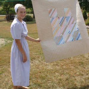 Puede incluir: Una persona joven con un vestido morado claro y un gorro blanco sostiene una colcha beige con un dise&ntilde;o de patchwork colorido. La colcha est&aacute; colgada de un tendedero.