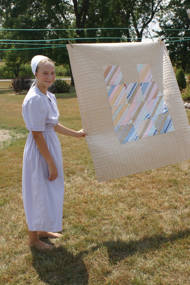 Puede incluir: Una persona joven con un vestido morado claro y un gorro blanco sostiene una colcha beige con un dise&ntilde;o de patchwork colorido. La colcha est&aacute; colgada de un tendedero.
