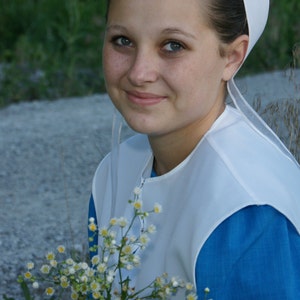 Pode incluir: Uma pessoa usando um gorro branco e um vestido azul, segurando um buqu&ecirc; de pequenas flores brancas e amarelas. A pessoa est&aacute; sorrindo, com um fundo de folhagem verde e um caminho de cascalho.