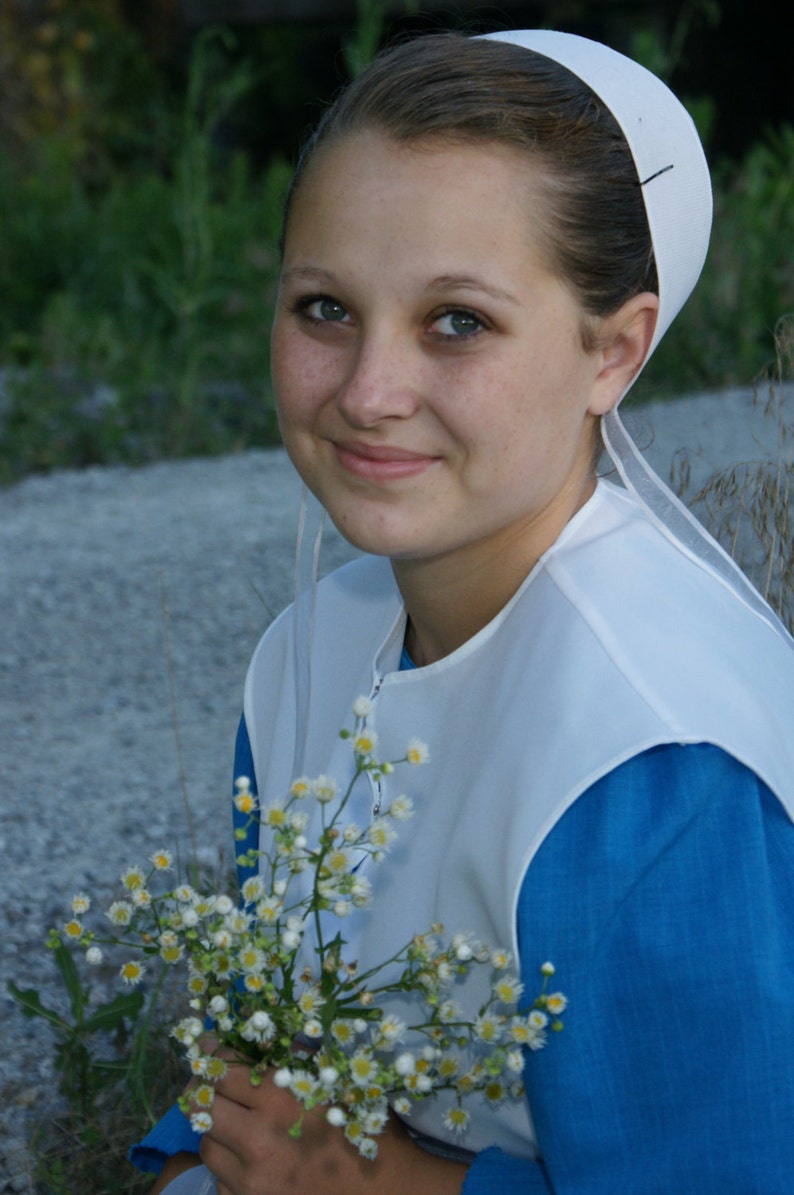 Pode incluir: Uma pessoa usando um gorro branco e um vestido azul, segurando um buqu&ecirc; de pequenas flores brancas e amarelas. A pessoa est&aacute; sorrindo, com um fundo de folhagem verde e um caminho de cascalho.