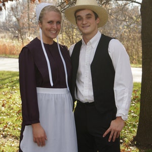 May include: A couple wearing traditional Amish clothing. The woman is wearing a long, dark purple dress with a white apron and a white cap. The man is wearing a white shirt, a black vest, and a straw hat. They are standing in a field with fall foliage.