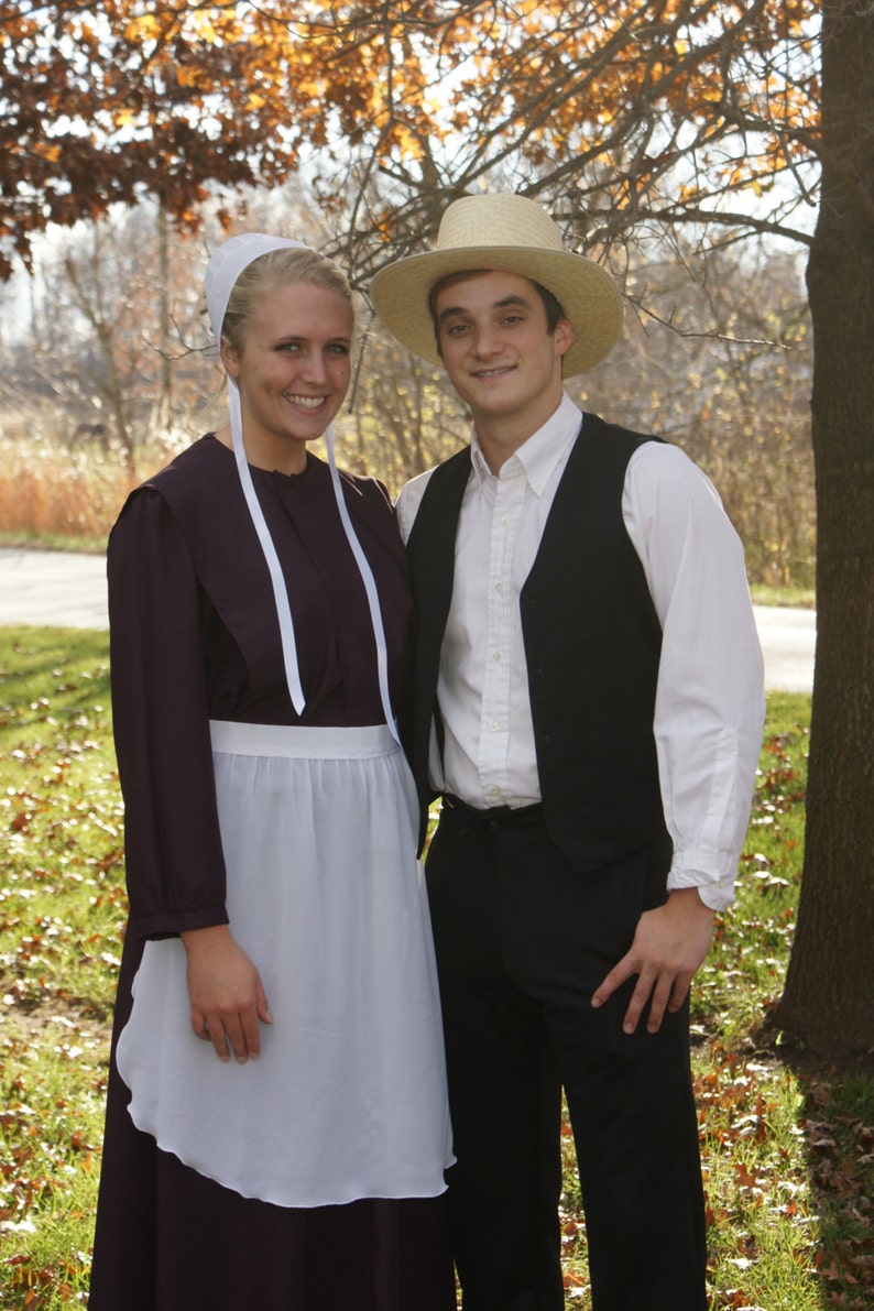 May include: A couple wearing traditional Amish clothing. The woman is wearing a long, dark purple dress with a white apron and a white cap. The man is wearing a white shirt, a black vest, and a straw hat. They are standing in a field with fall foliage.