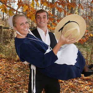 May include: A couple in an autumn setting. The woman is wearing a blue dress and a white bonnet. The man is wearing a black waistcoat and a white shirt. The woman is holding a straw hat.
