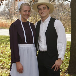 Puede incluir: Una joven con un vestido largo de color morado oscuro con un delantal blanco y un gorro blanco se encuentra junto a un joven que lleva una camisa blanca, un chaleco negro y un sombrero de paja. Ambos sonr&iacute;en y miran a la c&aacute;mara.