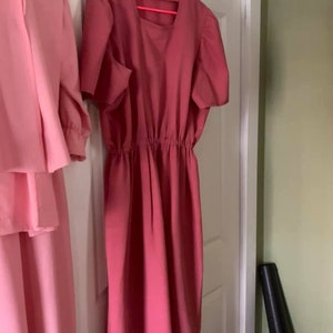 Puede incluir: Un vestido rosa con mangas cortas y una cintura fruncida. El vestido est&aacute; colgado en una percha rosa.