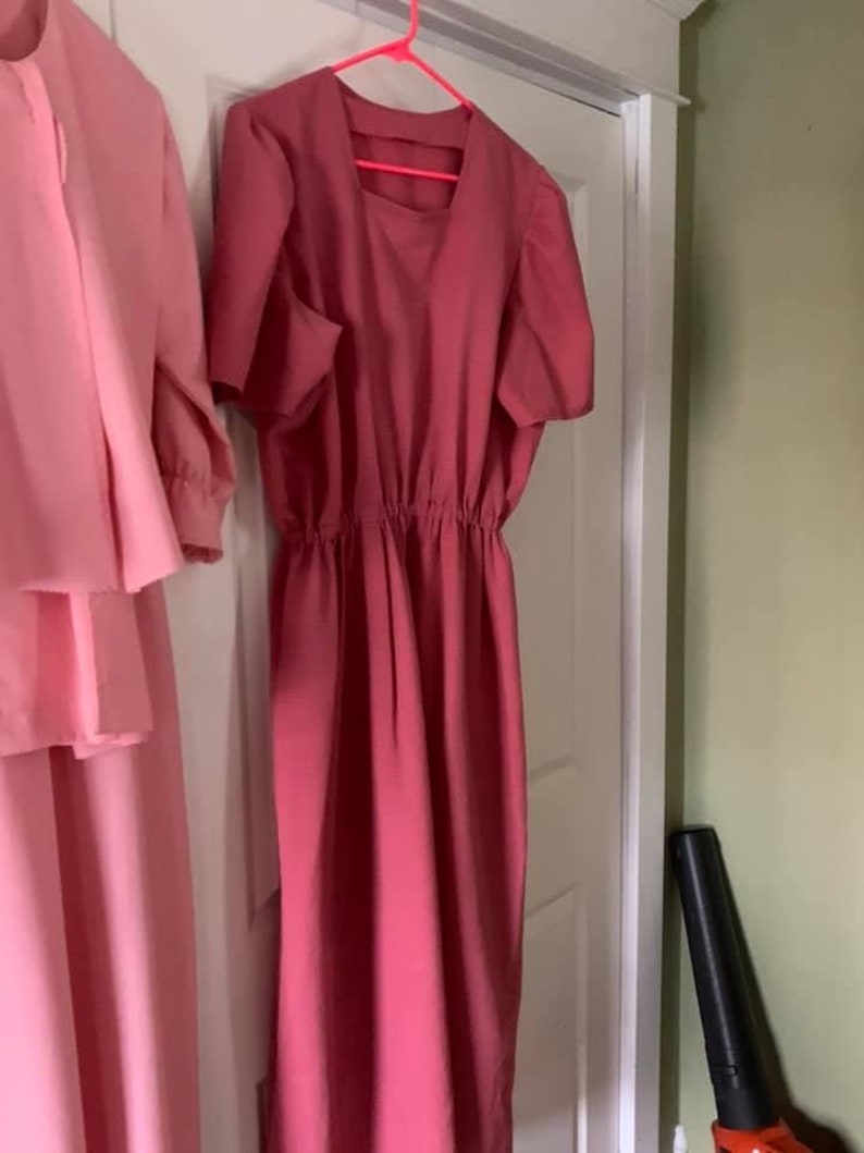 Puede incluir: Un vestido rosa con mangas cortas y una cintura fruncida. El vestido est&aacute; colgado en una percha rosa.