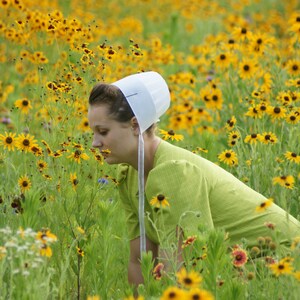 Pode incluir: Uma pessoa usando um gorro branco e um vestido verde, cercada por um campo de flores silvestres amarelas. A pessoa est&aacute; se inclinando, possivelmente cheirando as flores. A cena &eacute; ao ar livre em um dia ensolarado.