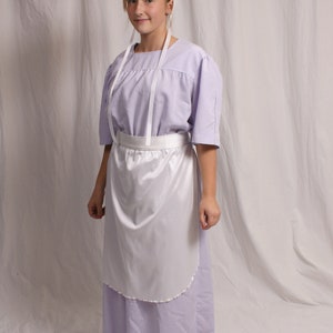 Puede incluir: Una persona joven que lleva un vestido lavanda con un delantal blanco y una cofia blanca. El vestido tiene una falda plisada y un corpi&ntilde;o sencillo. El delantal es simple, de forma recta con un dobladillo fruncido.