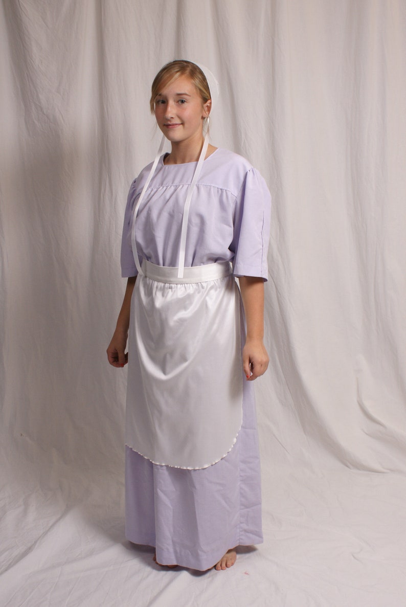 Puede incluir: Una persona joven que lleva un vestido lavanda con un delantal blanco y una cofia blanca. El vestido tiene una falda plisada y un corpi&ntilde;o sencillo. El delantal es simple, de forma recta con un dobladillo fruncido.