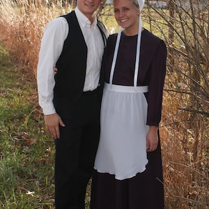 Puede incluir: Una pareja vestida con ropa tradicional Amish. El hombre lleva un sombrero de paja, una camisa blanca, un chaleco negro y pantalones negros. La mujer lleva una cofia blanca, un delantal blanco y un vestido largo oscuro.