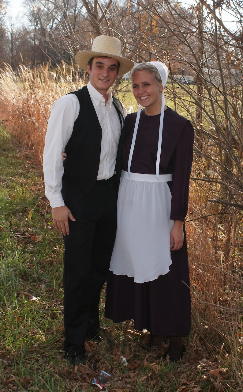 Puede incluir: Una pareja vestida con ropa tradicional Amish. El hombre lleva un sombrero de paja, una camisa blanca, un chaleco negro y pantalones negros. La mujer lleva una cofia blanca, un delantal blanco y un vestido largo oscuro.