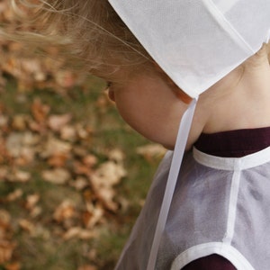 Pode incluir: Uma crian&ccedil;a usando um gorro branco e um avental transparente cinza claro sobre uma blusa bord&ocirc; escura. O gorro tem uma aba larga e amarra sob o queixo com uma fita branca. O fundo est&aacute; desfocado com folhagem de outono.