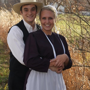 Puede incluir: Una pareja vestida con ropa tradicional Amish. El hombre lleva un sombrero de paja y un chaleco oscuro sobre una camisa blanca. La mujer lleva un vestido largo y oscuro con un delantal blanco y una cinta blanca alrededor del cuello.