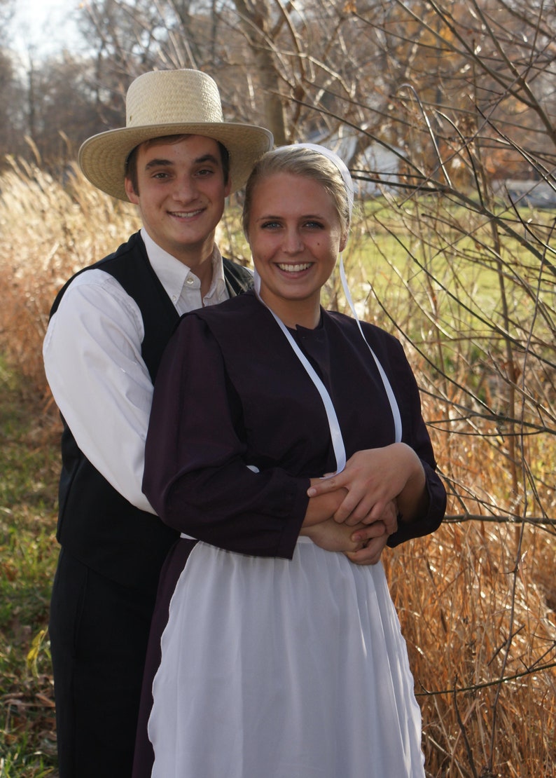 Puede incluir: Una pareja vestida con ropa tradicional Amish. El hombre lleva un sombrero de paja y un chaleco oscuro sobre una camisa blanca. La mujer lleva un vestido largo y oscuro con un delantal blanco y una cinta blanca alrededor del cuello.