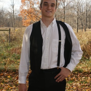 May include: A man wearing a white button-down shirt with a black vest and suspenders. He is standing in a field of fallen leaves.