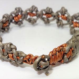 Puede incluir: Un brazalete de cuero trenzado con un cierre de plata. El brazalete está hecho de dos colores de cuero, un gris claro y un color cobre.