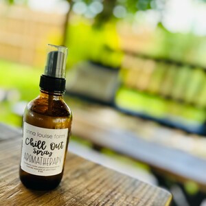 Puede incluir: Una botella de spray de aromaterapia de vidrio marrón con una bomba negra y una etiqueta blanca. La etiqueta dice "Chill Out Spray Aromatherapy" y "anna louise farm". La botella está sobre una superficie de madera.