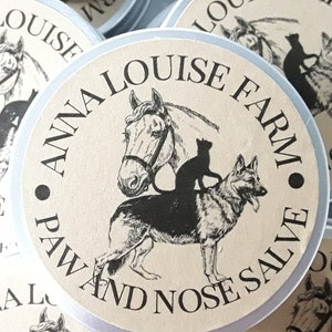 May include: A silver tin with a beige label that reads "Anna Louise Farm" and "Paw and Nose Salve". The label features a black and white illustration of a horse, a cat, and a dog.