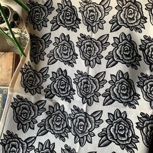 May include: Cream-coloured fabric featuring a repeating pattern of black rose illustrations. The roses are detailed with leaves and stems, creating a classic floral design. The fabric is hanging from a metal clip.