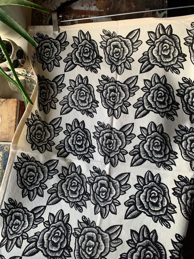 May include: Cream-coloured fabric featuring a repeating pattern of black rose illustrations. The roses are detailed with leaves and stems, creating a classic floral design. The fabric is hanging from a metal clip.