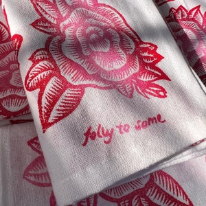 May include: White linen tea towels featuring a repeating pattern of a red rose and leaves. The towels have the handwritten text "jolly to some" in pink. The floral design is a bold, graphic print.